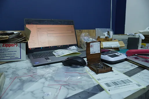 Paketleme masası: ekranda sipariş formu, açık markalı ahşap kutu, hassas terazi ve barkod etiketleri
